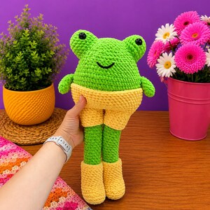 Op de afbeelding: Een handgemaakte, groene gehaakte kikkerpop met gele shorts en laarzen. De kikker heeft zwarte ogen en een lachend gezicht. De pop wordt vastgehouden voor een paarse achtergrond met een kleine plant in een gele pot en roze bloemen.