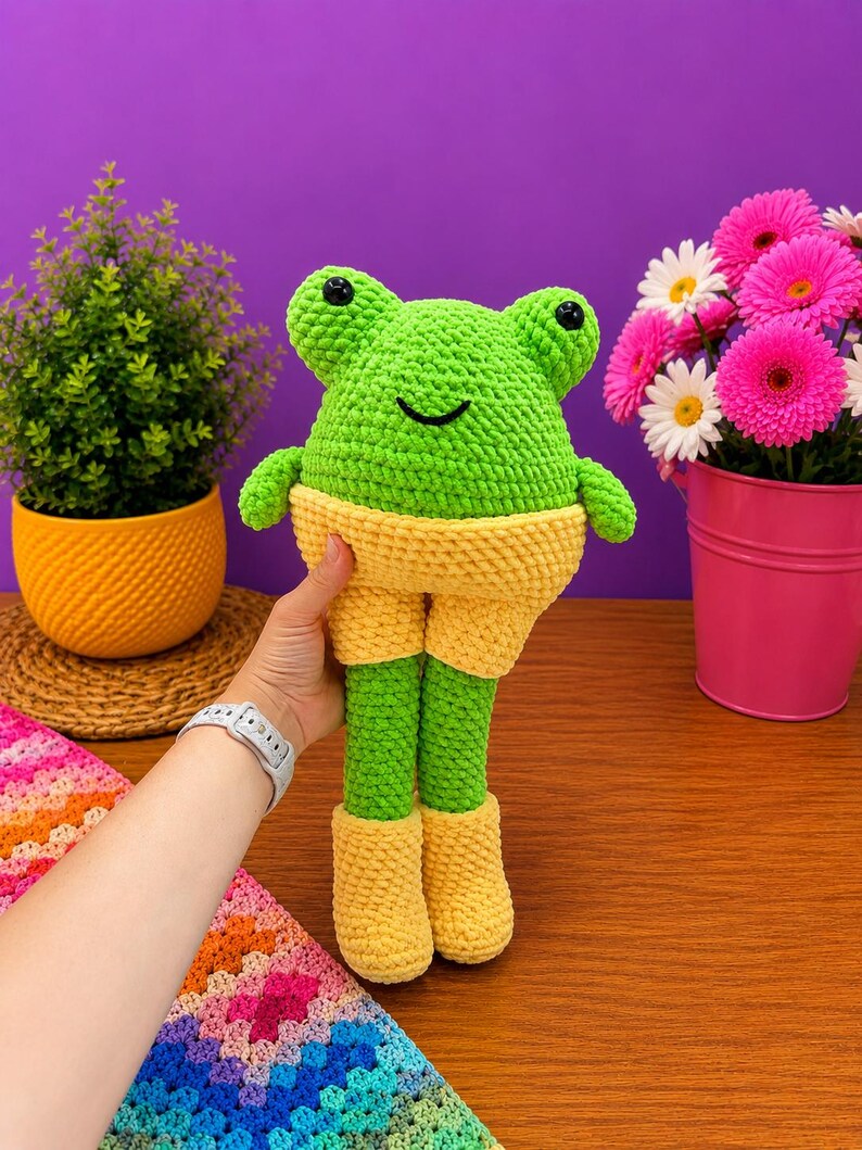 Op de afbeelding: Een handgemaakte, groene gehaakte kikkerpop met gele shorts en laarzen. De kikker heeft zwarte ogen en een lachend gezicht. De pop wordt vastgehouden voor een paarse achtergrond met een kleine plant in een gele pot en roze bloemen.
