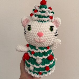 Pode incluir: Um gato de croch&ecirc; branco feito &agrave; m&atilde;o usando uma roupa de &aacute;rvore de Natal verde e branca, adornado com pompons vermelhos e enfeites prateados. O gato tem olhos verdes, um nariz rosa e bigodes pretos. O gato est&aacute; usando um chap&eacute;u de &aacute;rvore de Natal.