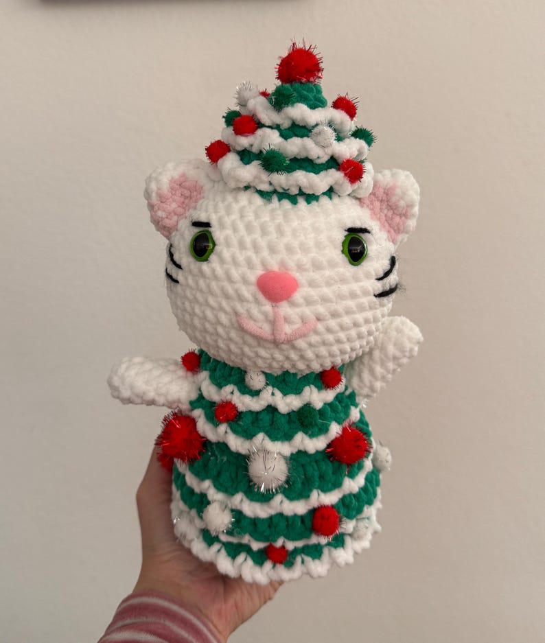 Pode incluir: Um gato de croch&ecirc; branco feito &agrave; m&atilde;o usando uma roupa de &aacute;rvore de Natal verde e branca, adornado com pompons vermelhos e enfeites prateados. O gato tem olhos verdes, um nariz rosa e bigodes pretos. O gato est&aacute; usando um chap&eacute;u de &aacute;rvore de Natal.