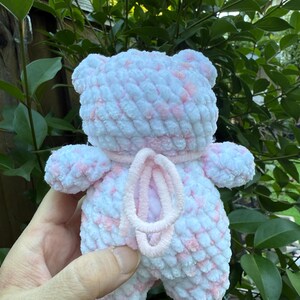 May include: A handmade, plush teddy bear in white and pink. The bear has a soft, textured appearance with a pink accent around the neck and a pink, looped design on the front. The bear is held against a backdrop of green foliage.