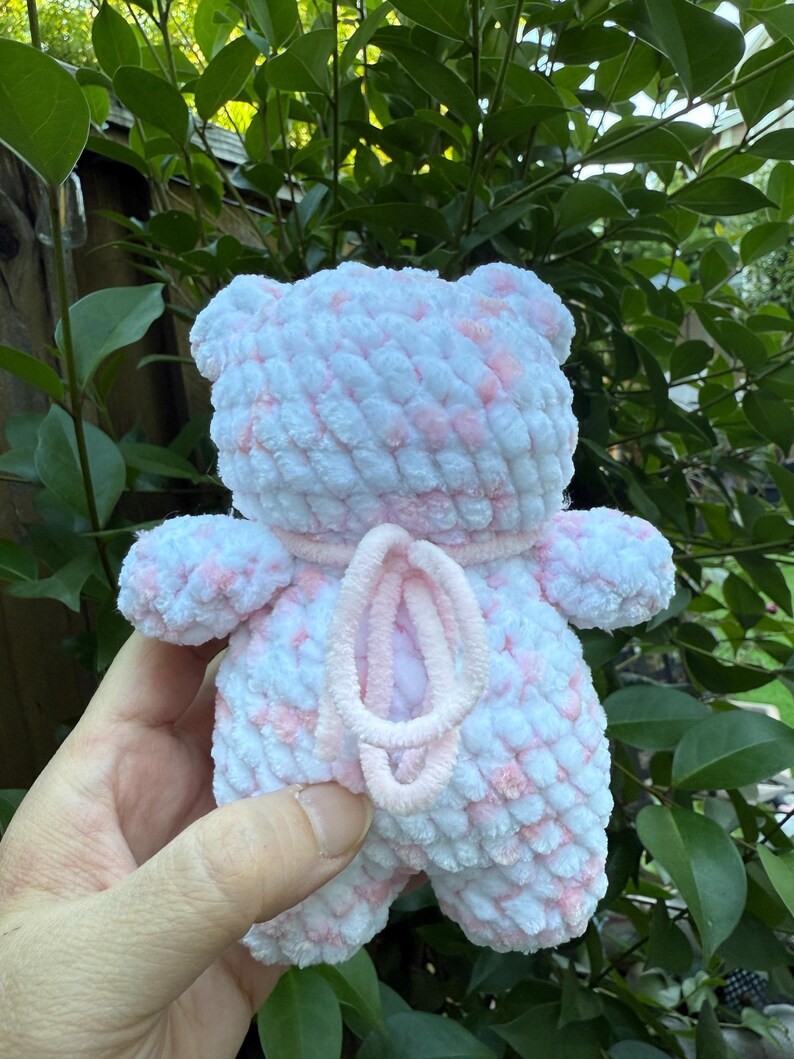 May include: A handmade, plush teddy bear in white and pink. The bear has a soft, textured appearance with a pink accent around the neck and a pink, looped design on the front. The bear is held against a backdrop of green foliage.