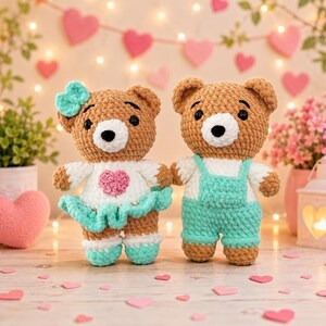K&ouml;nnte beinhalten: Zwei geh&auml;kelte Teddyb&auml;ren aus braunem Garn mit wei&szlig;en Schnauzen und schwarzen Augen. Der linke B&auml;r tr&auml;gt ein wei&szlig;es Kleid mit einem rosa Herz sowie einer t&uuml;rkisfarbenen Schleife, einem Rock und Schuhen. Der rechte B&auml;r tr&auml;gt wei&szlig;e &Auml;rmel und eine t&uuml;rkisfarbene Latzhose.