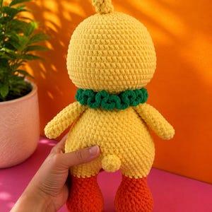 Op de afbeelding: Een handgemaakte gele gehaakte eendenpop met oranje voeten en een groene kraag. De pop heeft een afgeronde kop met een kleine pluk en kleine armen. De pop staat op een roze ondergrond met een oranje achtergrond.