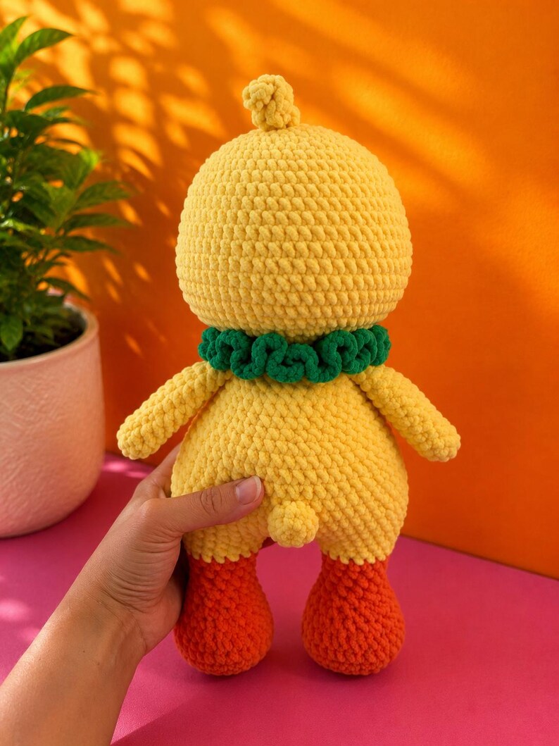 Op de afbeelding: Een handgemaakte gele gehaakte eendenpop met oranje voeten en een groene kraag. De pop heeft een afgeronde kop met een kleine pluk en kleine armen. De pop staat op een roze ondergrond met een oranje achtergrond.