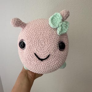 以下が含まれることがあります： 手作りのピンク色の編みぐるみぬいぐるみ。大きな黒い反射する目と笑顔の口があります。小さな薄いピンク色の耳と薄緑色のリボンが付いています。ぬいぐるみは丸くて柔らかいです。
