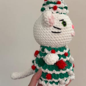 Pode incluir: Uma figura de gato branco feita &agrave; m&atilde;o em croch&ecirc;, usando um vestido e chap&eacute;u de &aacute;rvore de Natal verde e branco, adornada com pompons vermelhos. O gato tem olhos verdes, orelhas e nariz rosa e bigodes pretos. Uma decora&ccedil;&atilde;o festiva.