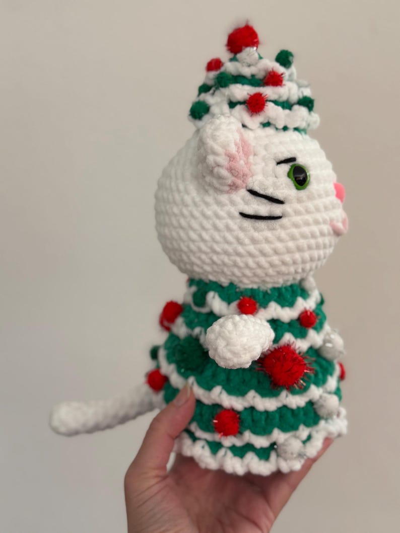 Pode incluir: Uma figura de gato branco feita &agrave; m&atilde;o em croch&ecirc;, usando um vestido e chap&eacute;u de &aacute;rvore de Natal verde e branco, adornada com pompons vermelhos. O gato tem olhos verdes, orelhas e nariz rosa e bigodes pretos. Uma decora&ccedil;&atilde;o festiva.
