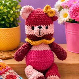 Op de afbeelding: Een gehaakte teddybeer met een kleurblokontwerp in tinten diep bordeaux, lichtroze en felmagenta. De beer heeft een gele gehaakte kraag om de nek, een kleine gele strik op het hoofd, zwarte plastic ogen en een geborduurde neus.