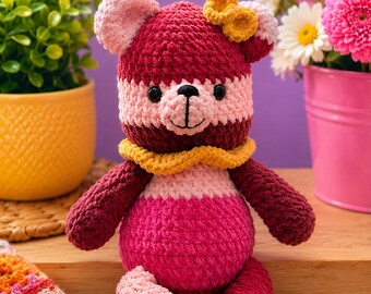 Peut inclure: Un ours en peluche crocheté avec un design en blocs de couleurs dans des tons de bordeaux profond, rose clair et magenta vif. L'ours porte un col crocheté jaune autour du cou, un petit nœud jaune sur la tête, des yeux en plastique noir et un nez brodé.