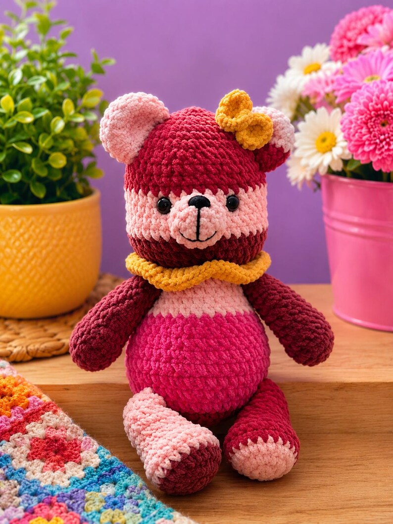 Op de afbeelding: Een gehaakte teddybeer met een kleurblokontwerp in tinten diep bordeaux, lichtroze en felmagenta. De beer heeft een gele gehaakte kraag om de nek, een kleine gele strik op het hoofd, zwarte plastic ogen en een geborduurde neus.
