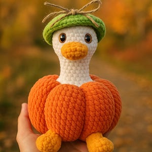 May include: A handmade crochet duck wearing a green hat with a bow. The duck has a white head, orange beak, and is wearing an orange pumpkin body. The duck's feet are also orange. The background is blurred with fall colors.