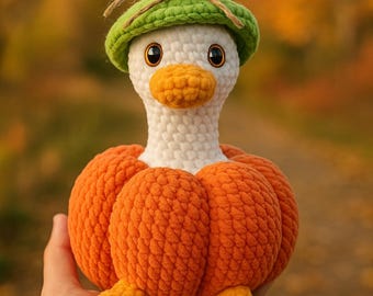 Op de afbeelding: Een handgemaakte gehaakte eend met een groene hoed met een strik. De eend heeft een witte kop, een oranje snavel en draagt een oranje pompoenlichaam. De voeten van de eend zijn ook oranje. De achtergrond is vervaagd met herfstkleuren.