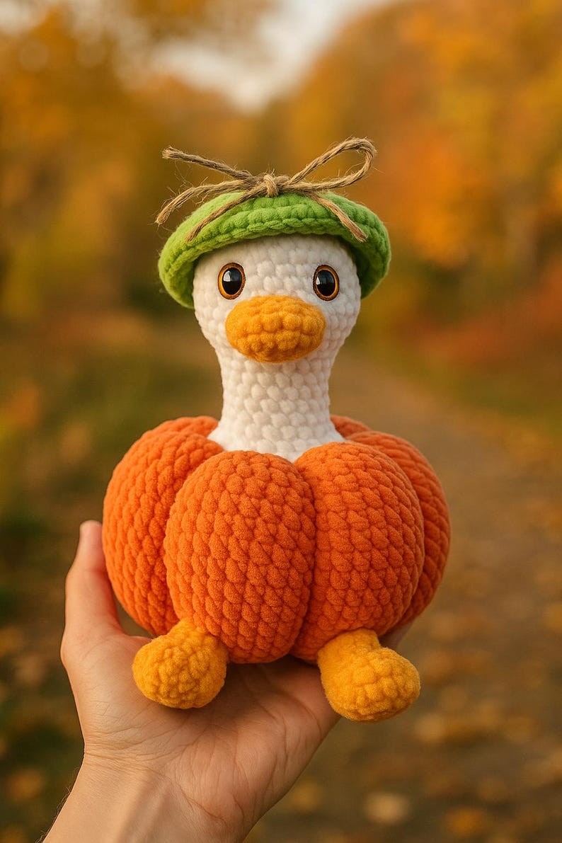 May include: A handmade crochet duck wearing a green hat with a bow. The duck has a white head, orange beak, and is wearing an orange pumpkin body. The duck's feet are also orange. The background is blurred with fall colors.