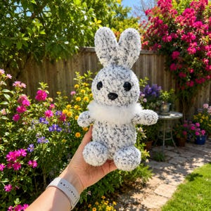 Peut inclure: Une peluche en forme de lapin avec un motif mouchet&eacute; blanc et gris. Le lapin a de grands yeux noirs, un nez noir et un col blanc duveteux. Il est pr&eacute;sent&eacute; devant un jardin fleuri.