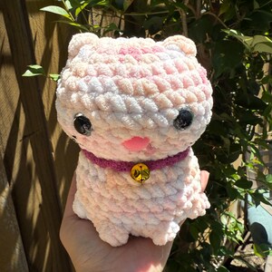 May include: A handmade, plush cat toy in shades of pink and white. The cat has black button eyes, a pink nose, and a purple collar with a gold bell. The toy is made of soft, textured yarn.
