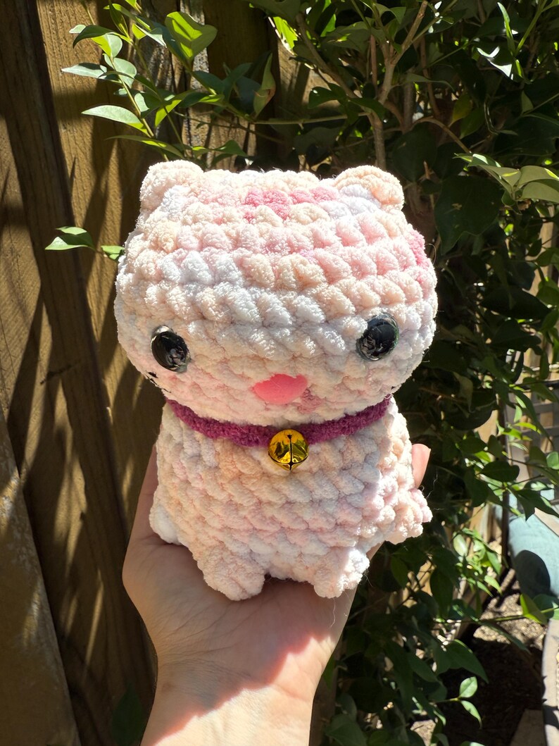 May include: A handmade, plush cat toy in shades of pink and white. The cat has black button eyes, a pink nose, and a purple collar with a gold bell. The toy is made of soft, textured yarn.