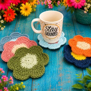 Op de afbeelding: Een witte keramische mok met de tekst Stay Grounded in zwarte letters, omringd door verschillende bloemvormige gehaakte onderzetters in blauw, roze, olijfgroen, oranje en marineblauw, elk met een contrasterende kleur in het midden.