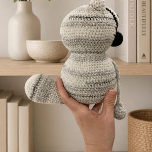May include: A small, round, crocheted stuffed animal featuring a textured, variegated yarn in shades of cream, gray, and black. The plush toy has a rounded head, a thick body, a curved tail, and a small, dark-colored nose visible on the side profile.