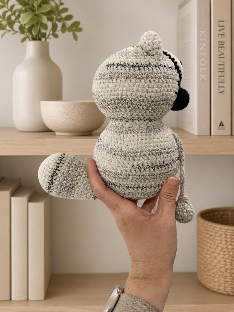 May include: A small, round, crocheted stuffed animal featuring a textured, variegated yarn in shades of cream, gray, and black. The plush toy has a rounded head, a thick body, a curved tail, and a small, dark-colored nose visible on the side profile.