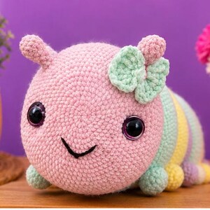 以下が含まれることがあります： パステルカラーの、手作りのカギ針編みで作られたイモムシのおもちゃ。ピンク色の頭には大きな黒い目、笑顔の口、ミントグリーンのリボンが付いています。体はパステルカラーのストライプです。手作りのアイテムです。