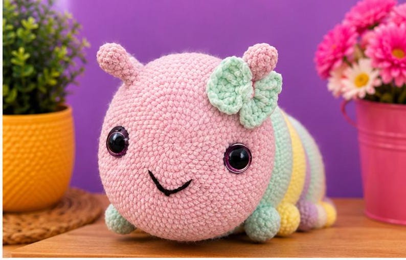 以下が含まれることがあります： パステルカラーの、手作りのカギ針編みで作られたイモムシのおもちゃ。ピンク色の頭には大きな黒い目、笑顔の口、ミントグリーンのリボンが付いています。体はパステルカラーのストライプです。手作りのアイテムです。