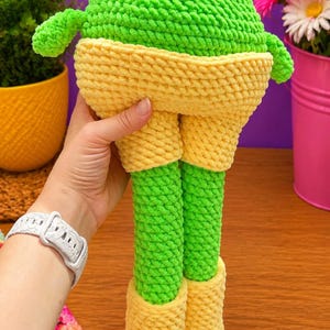 Op de afbeelding: Een handgemaakte gehaakte kikkerpop in heldergroen en geel. De kikker heeft een groene kop en poten, met gele shorts en schoenen. De pop is gemaakt van zacht, getextureerd garen. De pop wordt vastgehouden.