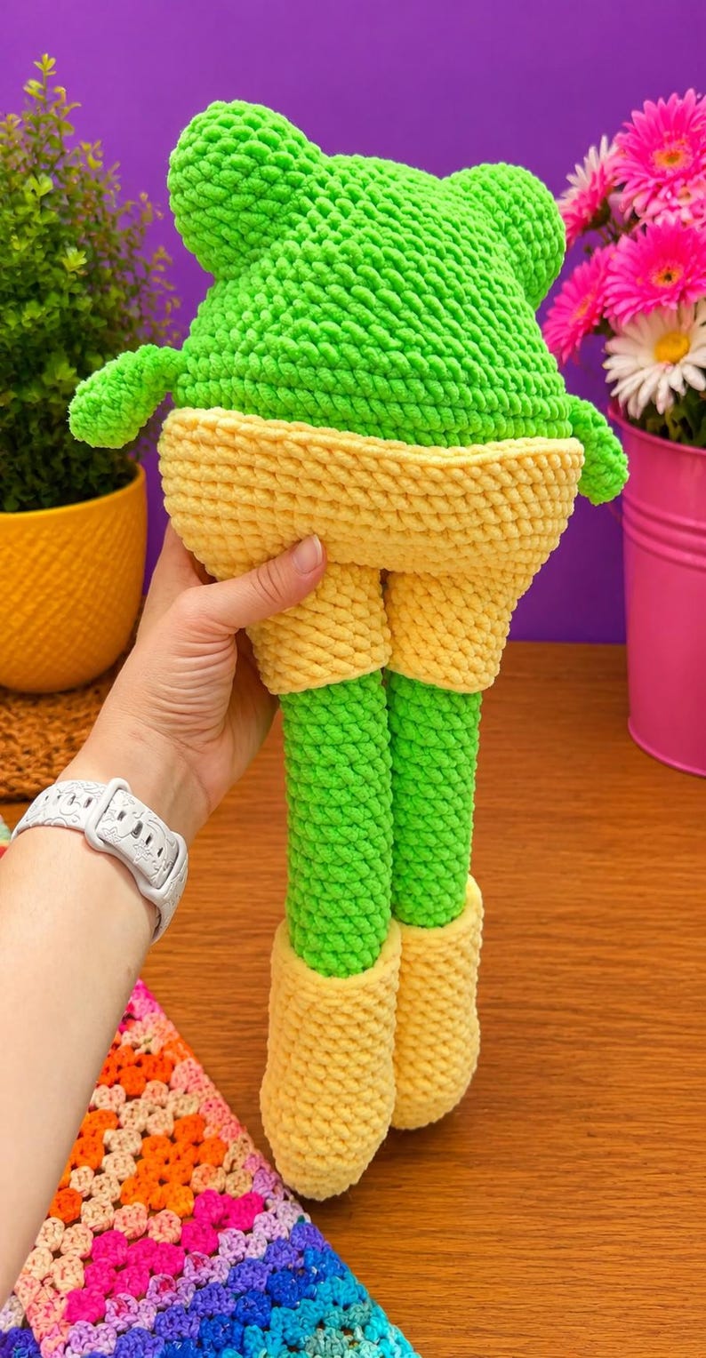 Op de afbeelding: Een handgemaakte gehaakte kikkerpop in heldergroen en geel. De kikker heeft een groene kop en poten, met gele shorts en schoenen. De pop is gemaakt van zacht, getextureerd garen. De pop wordt vastgehouden.