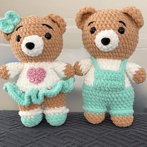 K&ouml;nnte beinhalten: Zwei handgefertigte H&auml;kel-Teddyb&auml;ren aus weichem beigem Garn mit wei&szlig;en Schnauzen und schwarzen Sicherheitsaugen. Der linke B&auml;r tr&auml;gt ein wei&szlig;es Oberteil mit einem rosa Herz, einen mintgr&uuml;nen Rock und eine passende Schleife. Der rechte B&auml;r tr&auml;gt eine Latzhose.