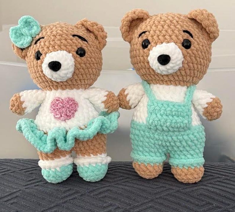 K&ouml;nnte beinhalten: Zwei handgefertigte H&auml;kel-Teddyb&auml;ren aus weichem beigem Garn mit wei&szlig;en Schnauzen und schwarzen Sicherheitsaugen. Der linke B&auml;r tr&auml;gt ein wei&szlig;es Oberteil mit einem rosa Herz, einen mintgr&uuml;nen Rock und eine passende Schleife. Der rechte B&auml;r tr&auml;gt eine Latzhose.