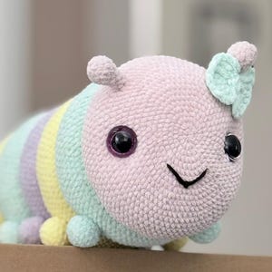 以下が含まれることがあります： パステルカラーの手編みかぎ針編みの毛虫のおもちゃ。毛虫は、大きな黒い目と笑顔の口を持つピンク色の頭をしています。体は青、黄色、紫の色合いでストライプになっています。おもちゃは柔らかく、抱きしめたくなるように設計されています。