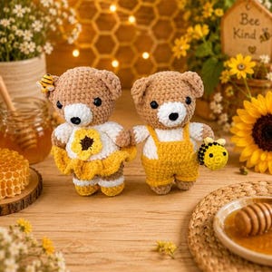 K&ouml;nnte beinhalten: Zwei geh&auml;kelte Teddyb&auml;ren stehen nebeneinander. Der linke B&auml;r tr&auml;gt ein wei&szlig;es Oberteil mit einer gelben Sonnenblume und einen gelben Rock sowie eine kleine Biene am Ohr. Der rechte B&auml;r tr&auml;gt eine gelbe Latzhose und h&auml;lt eine kleine geh&auml;kelte Biene.