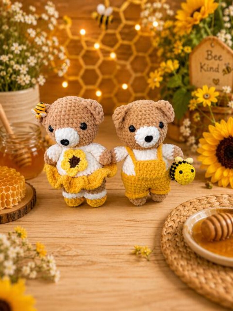 K&ouml;nnte beinhalten: Zwei geh&auml;kelte Teddyb&auml;ren stehen nebeneinander. Der linke B&auml;r tr&auml;gt ein wei&szlig;es Oberteil mit einer gelben Sonnenblume und einen gelben Rock sowie eine kleine Biene am Ohr. Der rechte B&auml;r tr&auml;gt eine gelbe Latzhose und h&auml;lt eine kleine geh&auml;kelte Biene.