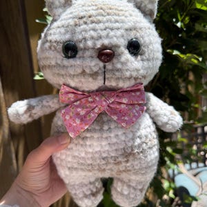 Pu&ograve; includere: Una bambola a forma di gatto all'uncinetto fatta a mano, nei toni del grigio e del bianco, con occhi neri e un papillon rosa floreale. Il peluche ha un corpo arrotondato e piccole braccia e gambe, ideale per un giocattolo per bambini.