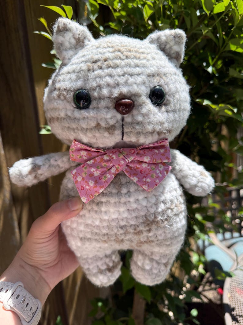 Pu&ograve; includere: Una bambola a forma di gatto all'uncinetto fatta a mano, nei toni del grigio e del bianco, con occhi neri e un papillon rosa floreale. Il peluche ha un corpo arrotondato e piccole braccia e gambe, ideale per un giocattolo per bambini.