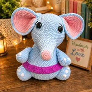 Pu&ograve; includere: Un giocattolo a forma di elefante realizzato all'uncinetto con filato azzurro, grandi orecchie con sezioni interne rosa, naso rosa e una fascia viola intorno alla vita. L'elefante ha occhi scuri e lucidi e piccole macchie bianche su piedi e braccia.