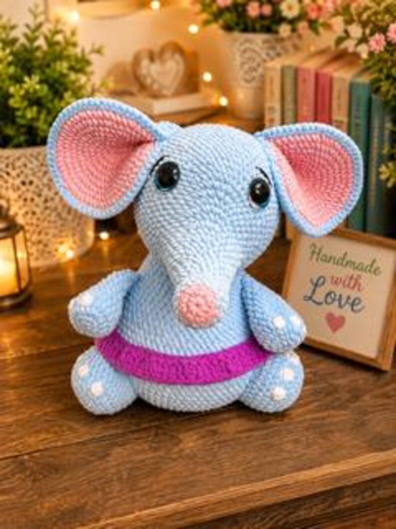 Pu&ograve; includere: Un giocattolo a forma di elefante realizzato all'uncinetto con filato azzurro, grandi orecchie con sezioni interne rosa, naso rosa e una fascia viola intorno alla vita. L'elefante ha occhi scuri e lucidi e piccole macchie bianche su piedi e braccia.