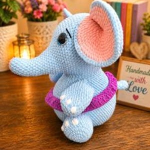 Pu&ograve; includere: Un peluche a forma di elefante realizzato all'uncinetto in azzurro, con grandi orecchie interne rosa, una fascia in vita viola scuro e dettagli ricamati bianchi sui piedi. L'elefante ha un corpo arrotondato, una proboscide e occhi neri.