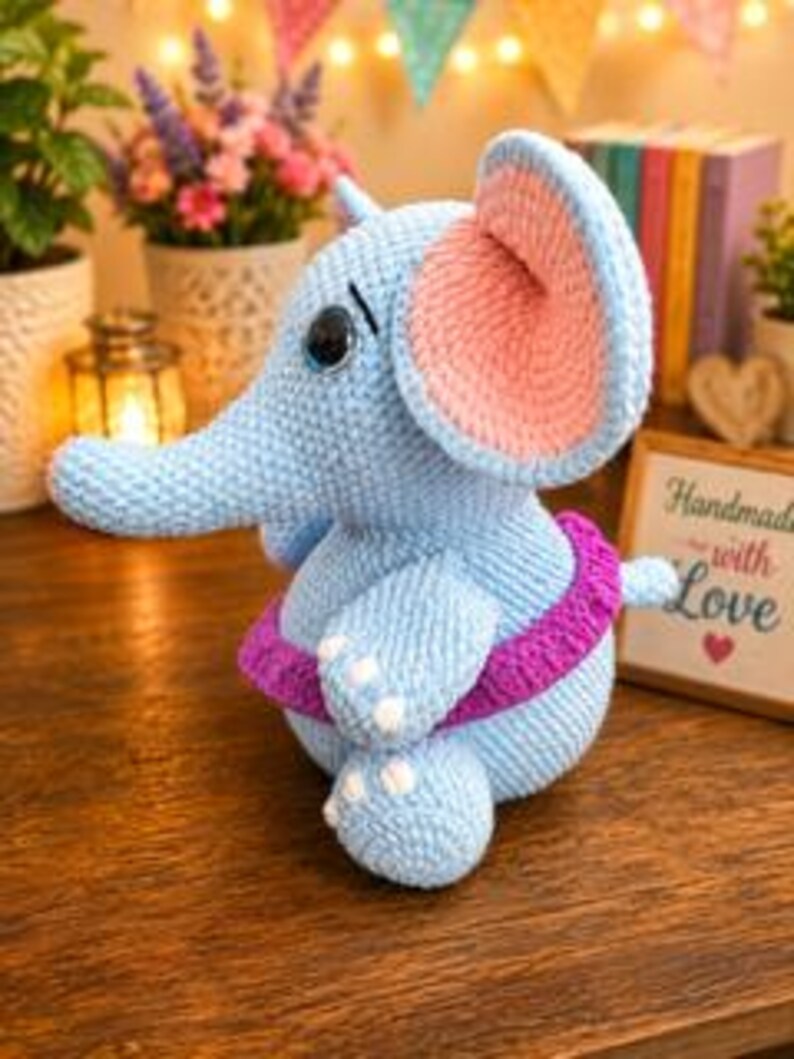 Pu&ograve; includere: Un peluche a forma di elefante realizzato all'uncinetto in azzurro, con grandi orecchie interne rosa, una fascia in vita viola scuro e dettagli ricamati bianchi sui piedi. L'elefante ha un corpo arrotondato, una proboscide e occhi neri.