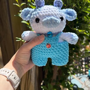 K&ouml;nnte beinhalten: Handgefertigtes geh&auml;keltes Kuh-Pl&uuml;schtier in Blau- und Wei&szlig;t&ouml;nen. Das Pl&uuml;schtier hat einen hellblauen Overall, kleine Ohren und einen Glockenhalsband. Ein roter Marienk&auml;fer-Knopf befindet sich auf dem Overall.