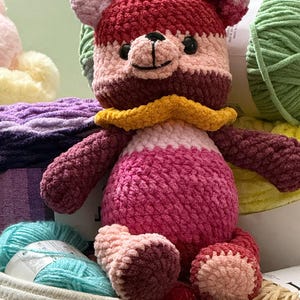 Op de afbeelding: Een gehaakte teddybeer gemaakt van pluche garen in tinten donkerroze, lichtroze en bordeauxrood. De beer heeft een gele ruchekraag, een gele strik op het hoofd en zwarte plastic ogen. Het zit tussen verschillende bollen garen in.