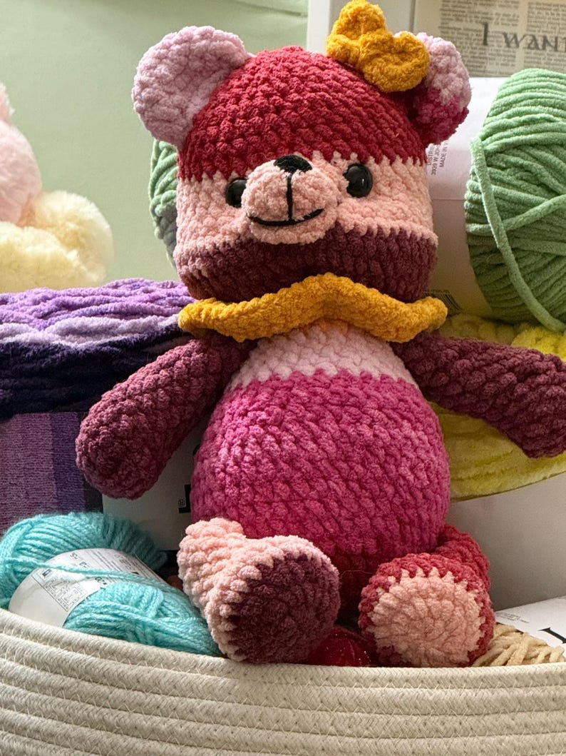 Op de afbeelding: Een gehaakte teddybeer gemaakt van pluche garen in tinten donkerroze, lichtroze en bordeauxrood. De beer heeft een gele ruchekraag, een gele strik op het hoofd en zwarte plastic ogen. Het zit tussen verschillende bollen garen in.