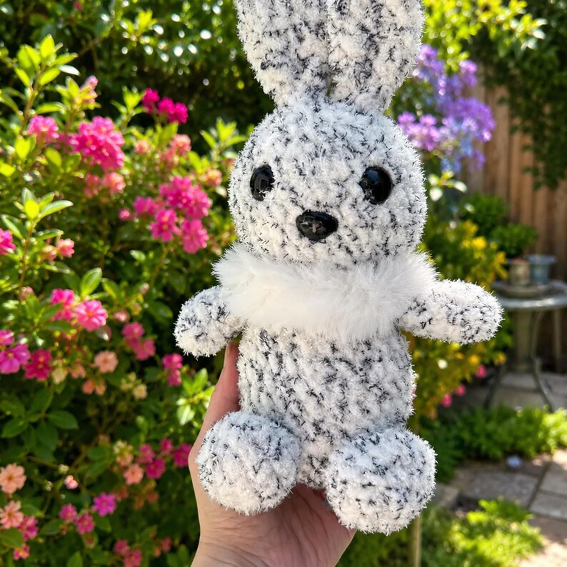 Peut inclure: Un lapin en peluche fait &agrave; la main, avec une fourrure tachet&eacute;e noire et blanche, de grands yeux noirs et un col blanc duveteux. Le lapin mesure environ 20 cm de haut et a une expression amicale. Le fond pr&eacute;sente un feuillage rose et vert vif.
