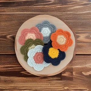 Op de afbeelding: Een verzameling van zes bloemvormige gehaakte onderzetters in diverse kleuren, waaronder oudroze, olijfgroen, lichtblauw, donkerblauw en oranje. Elke onderzetter heeft een contrasterend rond midden en geschulpte randen in een groepje.