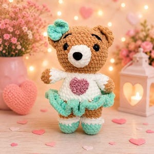 K&ouml;nnte beinhalten: Ein geh&auml;kelter Teddyb&auml;r mit braunem Fell, wei&szlig;er Schnauze und schwarzen Augen. Er tr&auml;gt ein wei&szlig;es Oberteil mit einem strukturierten rosa Herz, einen mintgr&uuml;nen R&uuml;schenrock, passende mintgr&uuml;ne Schuhe und eine mintgr&uuml;ne Schleife am Kopf.