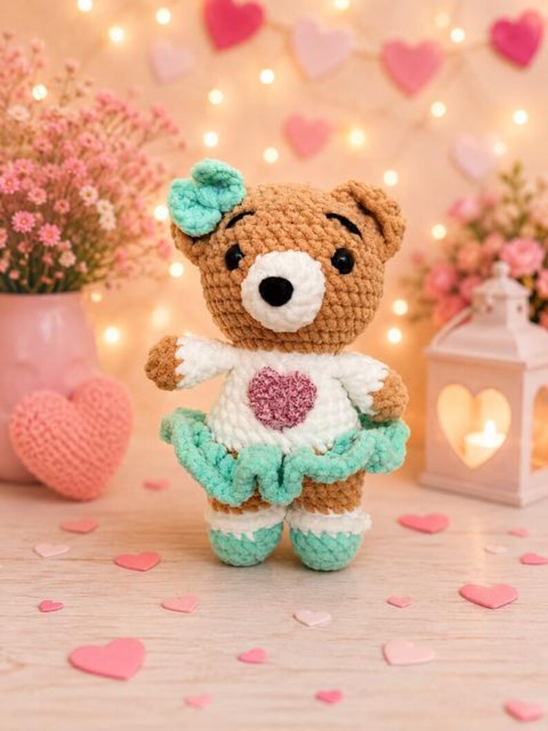 K&ouml;nnte beinhalten: Ein geh&auml;kelter Teddyb&auml;r mit braunem Fell, wei&szlig;er Schnauze und schwarzen Augen. Er tr&auml;gt ein wei&szlig;es Oberteil mit einem strukturierten rosa Herz, einen mintgr&uuml;nen R&uuml;schenrock, passende mintgr&uuml;ne Schuhe und eine mintgr&uuml;ne Schleife am Kopf.