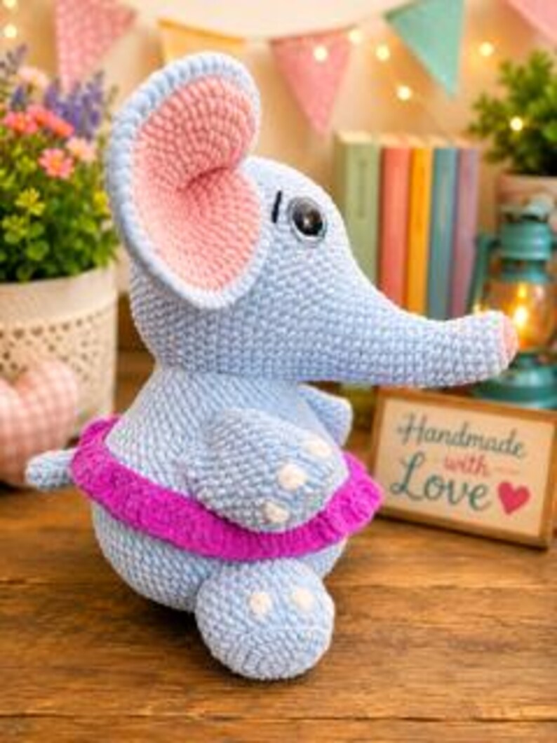 Pu&ograve; includere: Un peluche a forma di elefante lavorato all'uncinetto in azzurro, con grandi orecchie interne rosa, un occhio scuro e un vivace tut&ugrave; magenta intorno alla vita. L'elefante presenta piccoli dettagli bianchi sui piedi e una proboscide rivolta a destra.