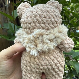 Peut inclure: Un ours en crochet beige fait &agrave; la main, avec un col en fourrure marron clair. L'ours est tenu dans la main d'une personne, avec un feuillage vert en arri&egrave;re-plan. Le corps textur&eacute; et les petites oreilles de l'ours sont visibles.