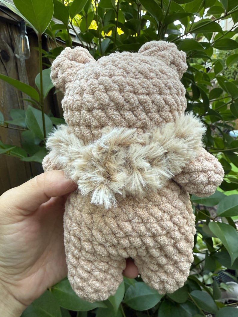 Peut inclure: Un ours en crochet beige fait &agrave; la main, avec un col en fourrure marron clair. L'ours est tenu dans la main d'une personne, avec un feuillage vert en arri&egrave;re-plan. Le corps textur&eacute; et les petites oreilles de l'ours sont visibles.