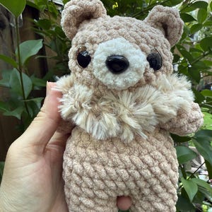 Peut inclure: Un ours en crochet marron clair avec un museau blanc duveteux et un nez noir. L'ours a de grands yeux noirs et de petites oreilles arrondies. L'ours est tenu dans la main d'une personne sur un fond de feuilles vertes.